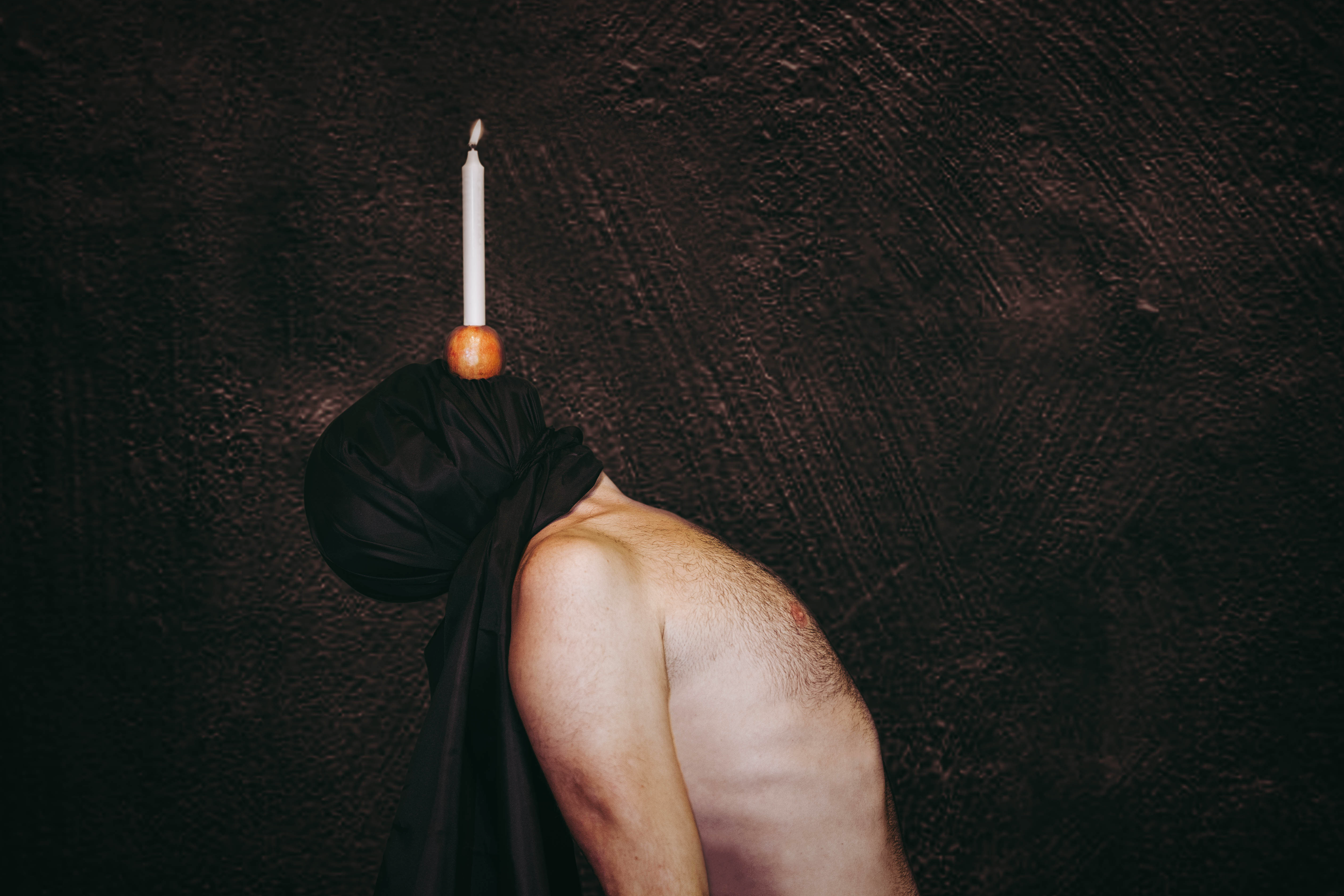 A white queer body is positioned sideways on in the centre of the image. Their naked torso and head is bending backwards, there is a black piece of fabric covering the artist’s face, a candle resting in an apple is being held on top of the fabric in the artists mouth.