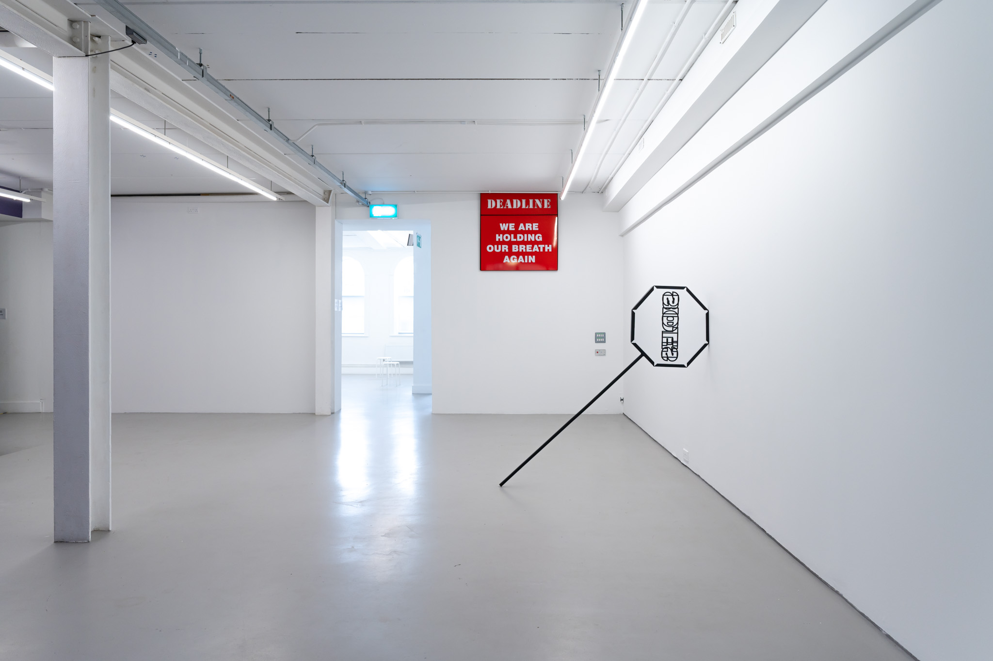 A black circular picket sign is resting on a wall to the right in a white room. On the back wall is a square red sign with white writing, it reads; deadline, we are holding our breath again.